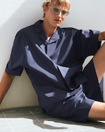 Placeholder for: Person with blond hair and sunglasses wearing a loose-fitting navy outfit sits against a white wall on tiled flooring.