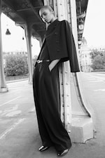 Placeholder for: A person in a black outfit leans against a metal bridge pillar, with a cityscape and trees in the background. The scene is elegant and monochrome.