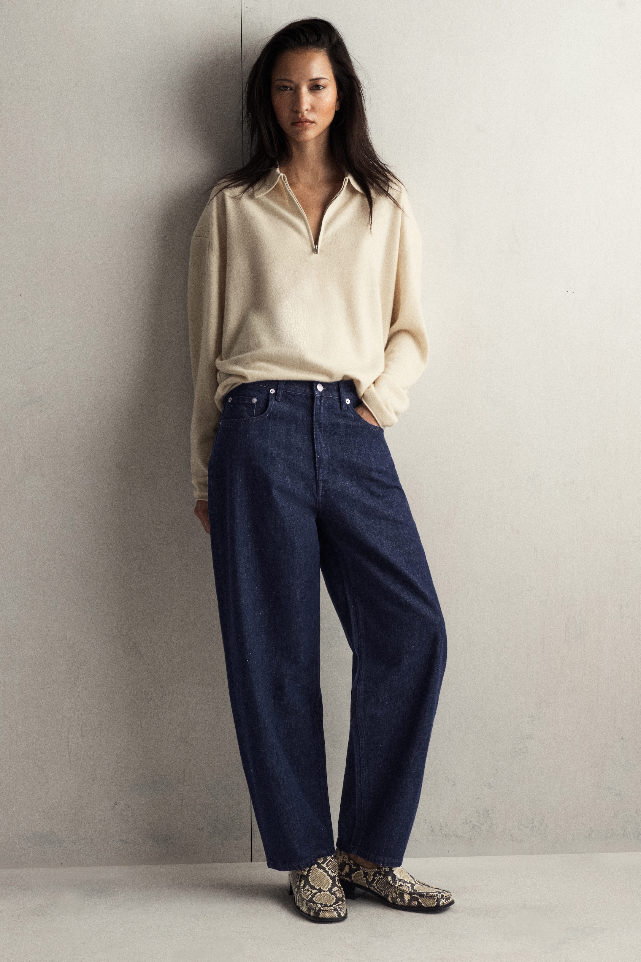 Woman in a beige sweater and wide-leg jeans, standing against a pale wall, wearing snake-patterned boots.