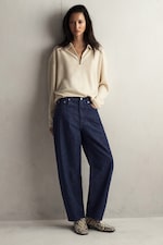 Placeholder for: Woman in a beige sweater and wide-leg jeans, standing against a pale wall, wearing snake-patterned boots.