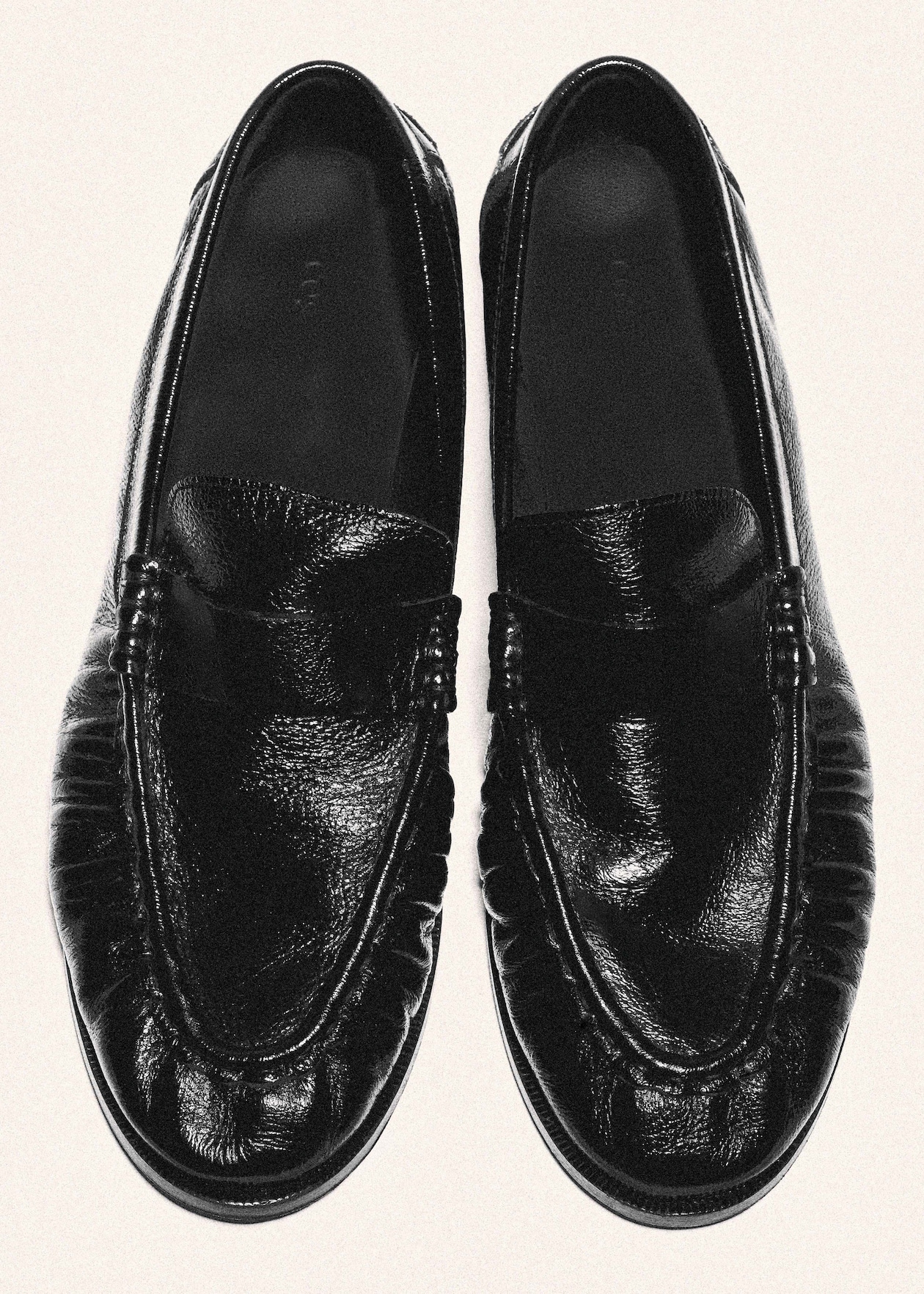 A pair of shiny black leather slip-on loafers with pleated detailing on the uppers, viewed from above on a light background.