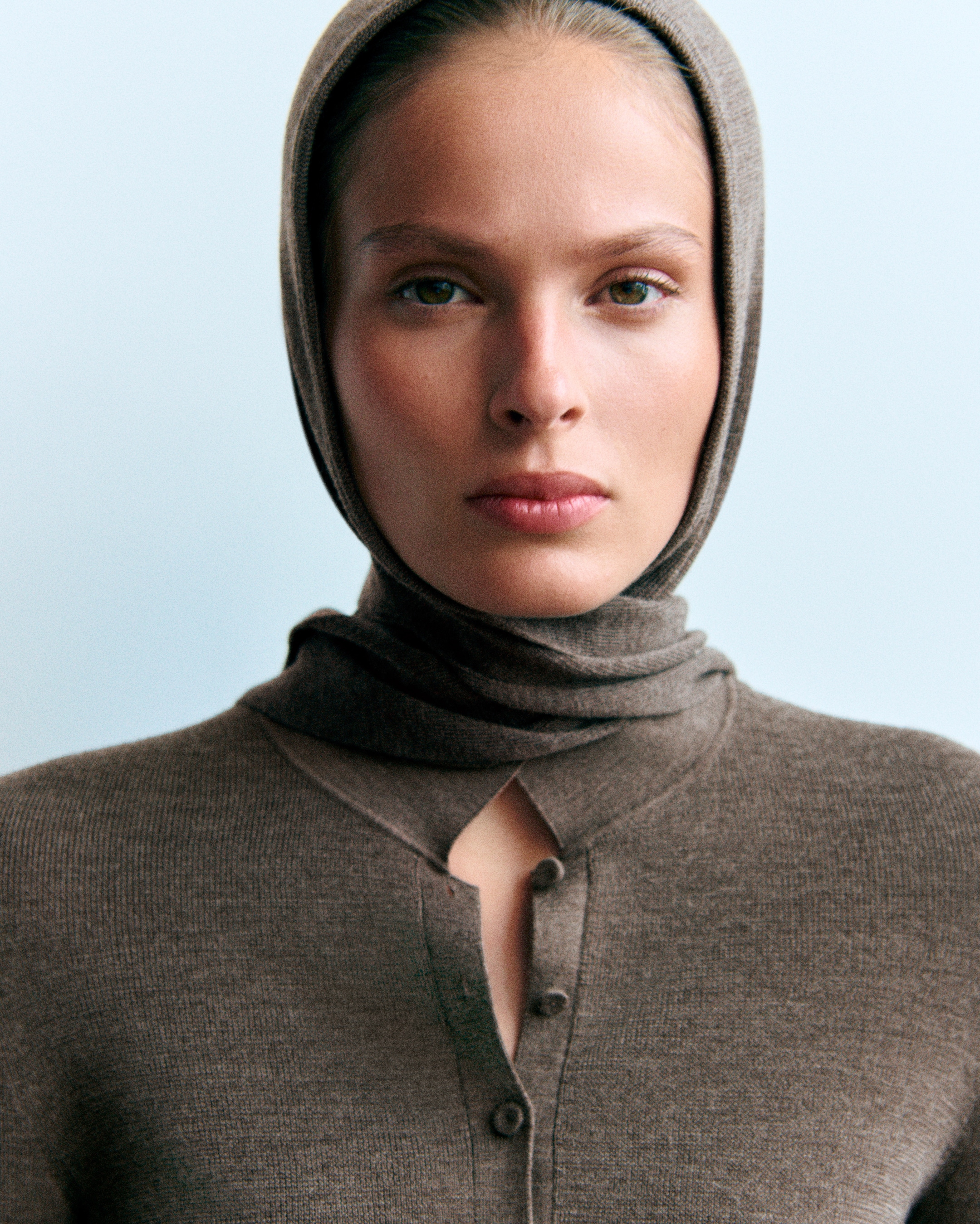 Person gazing forward, wearing a brown headscarf and sweater with a light blue background.