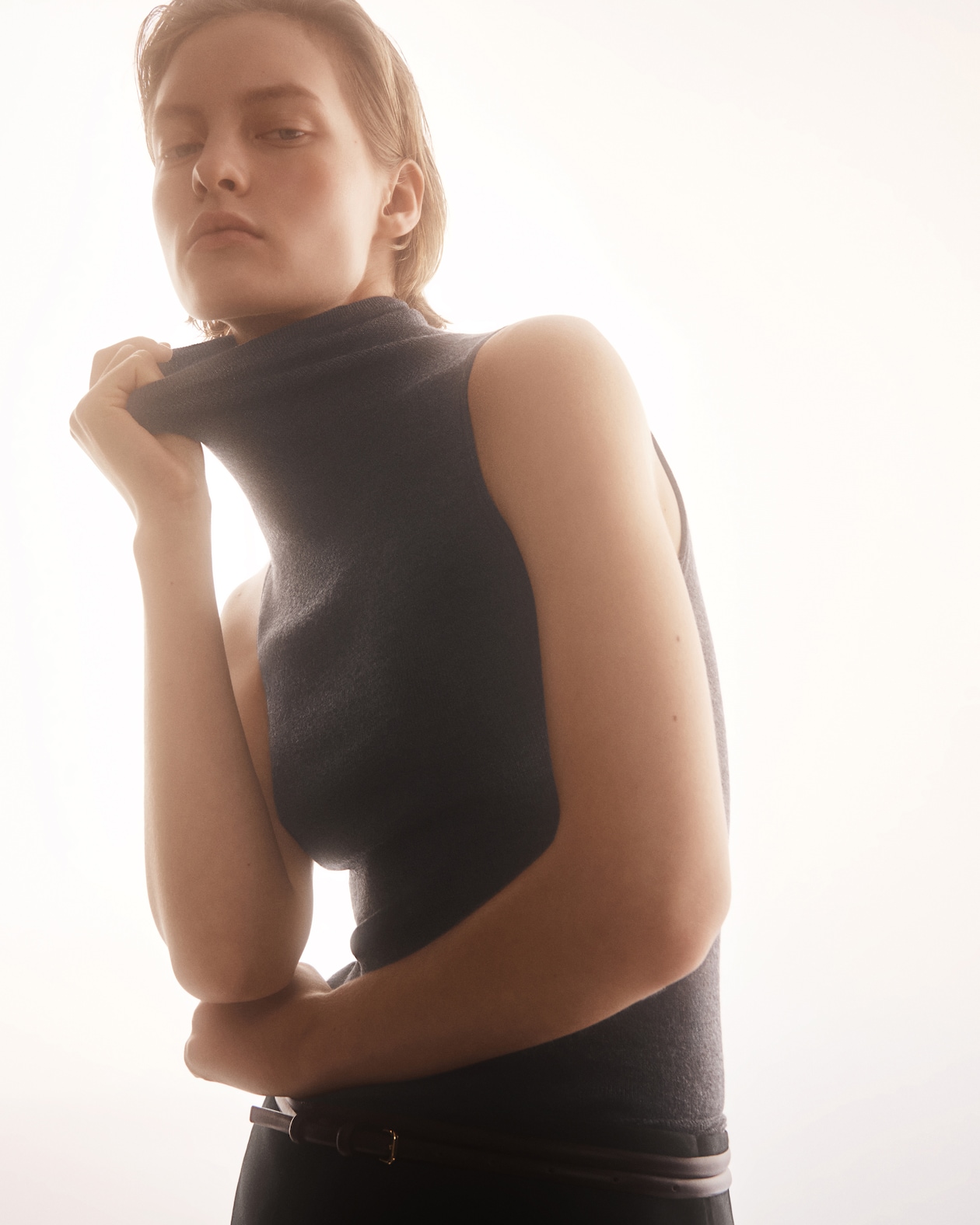 Person wearing a sleeveless turtleneck top poses against a soft, backlit background, with one hand adjusting the collar.