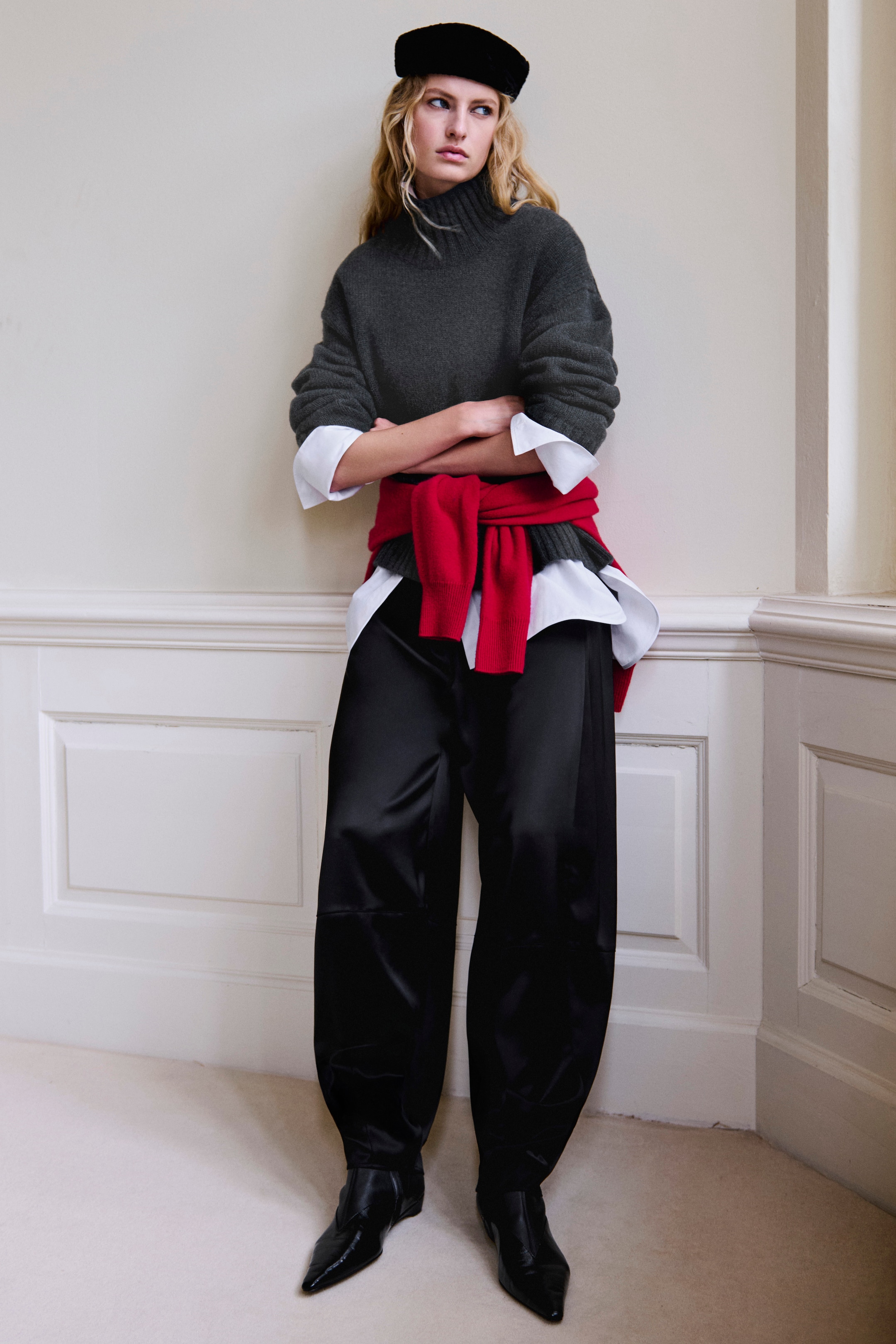 A person wearing a gray sweater, red scarf, and black pants stands against a white paneled wall, looking to the side with arms crossed.