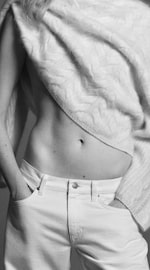 Placeholder for: Person in a cable-knit sweater partially lifted, revealing a toned midriff, and wearing white jeans with hands in pockets; shown in black and white.