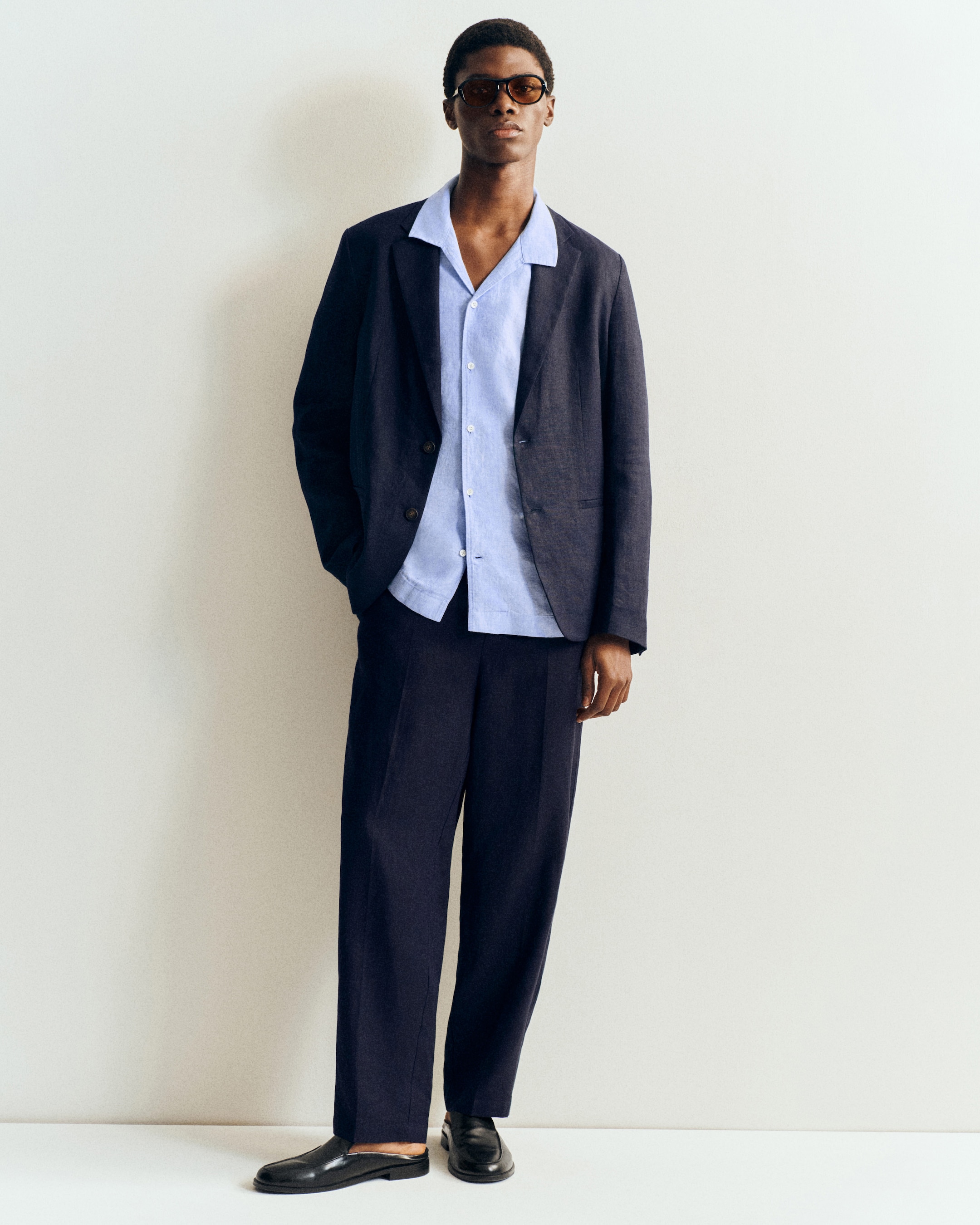 Man in sunglasses wearing a dark suit jacket, light blue shirt, and matching trousers, stands against a plain white background.