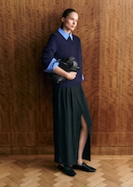 Placeholder for: A person in a purple sweater with blue cuffs and collar, holding a black bag. They wear a long skirt and black shoes, standing against a wooden wall.