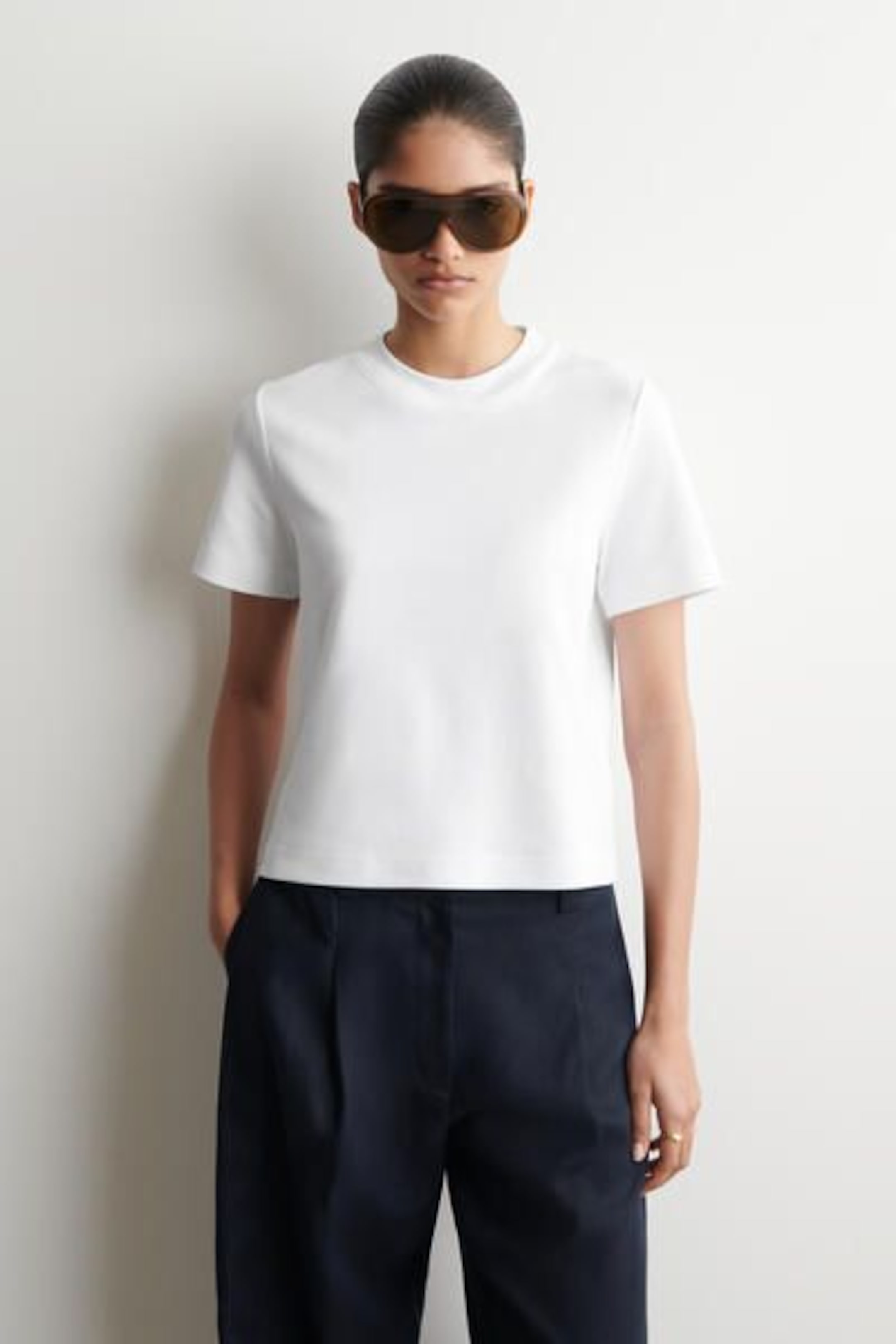 Person wearing a white T-shirt, dark pants, and sunglasses, standing against a plain background with hands in pockets.