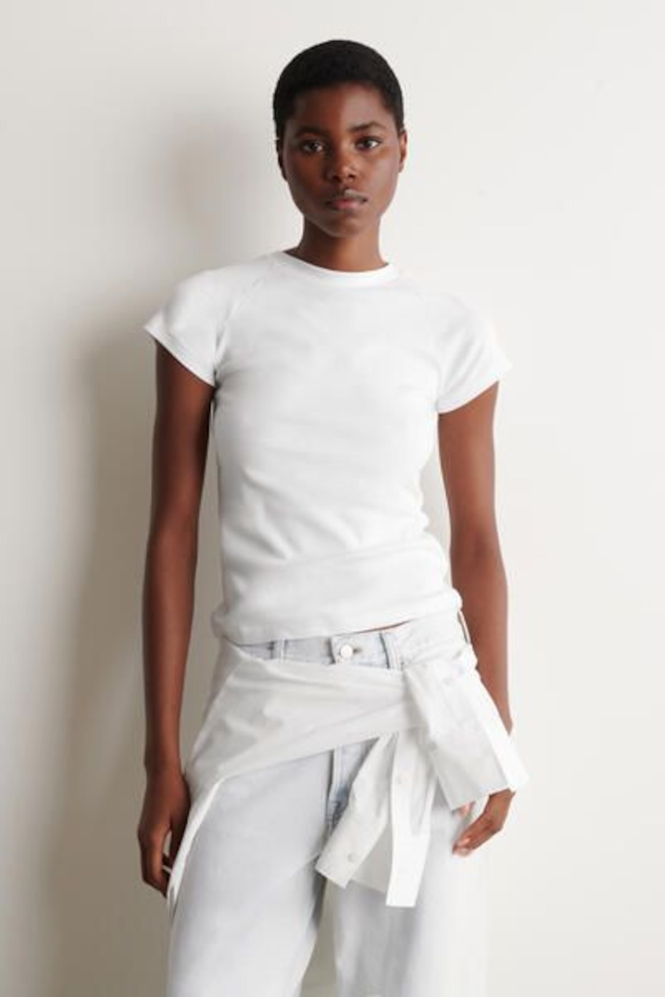 A person wearing a fitted white t-shirt and light-wash jeans with a tied white shirt around the waist, standing against a plain background.