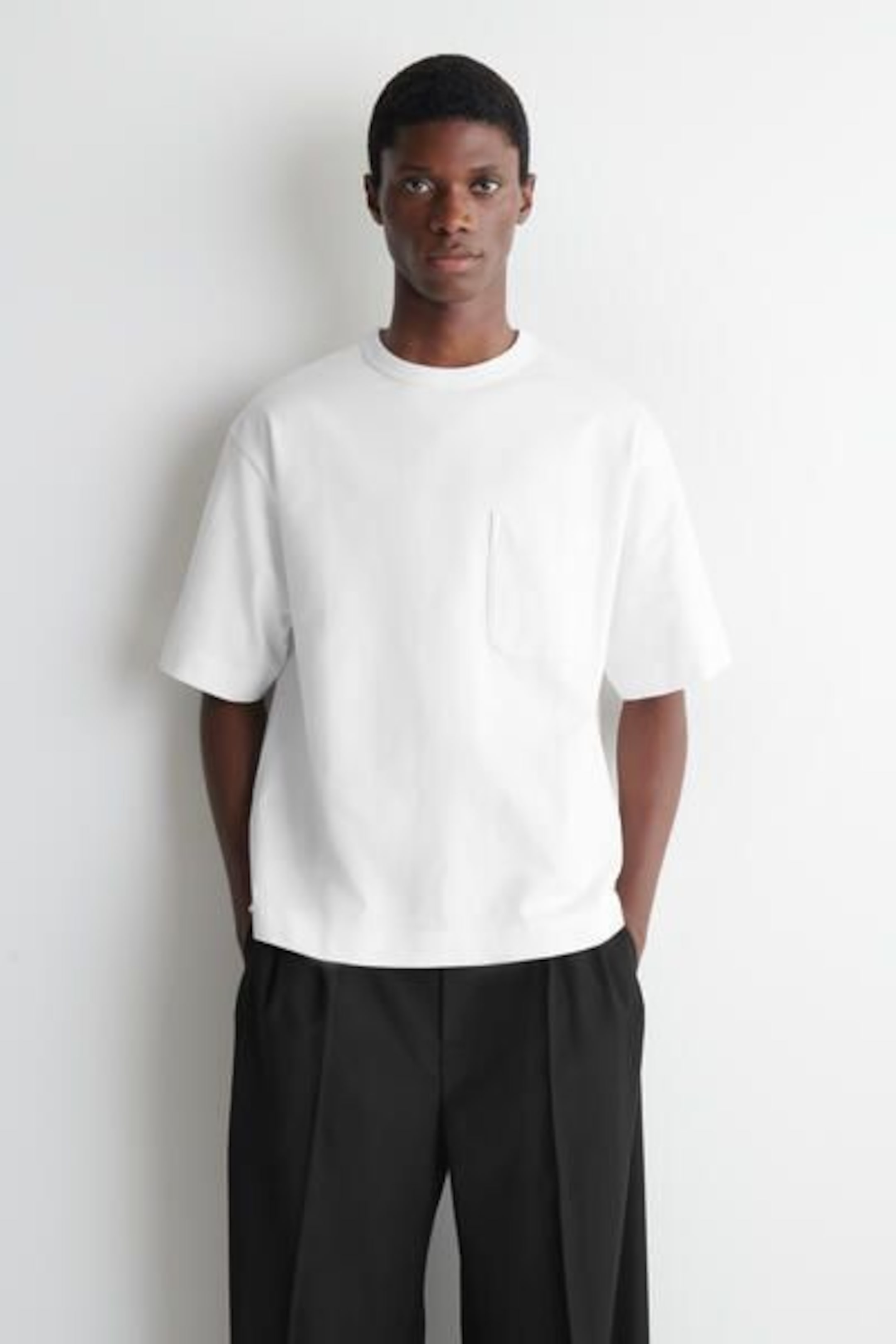 Person wearing a white T-shirt and black pants, standing against a white background with hands in pockets.