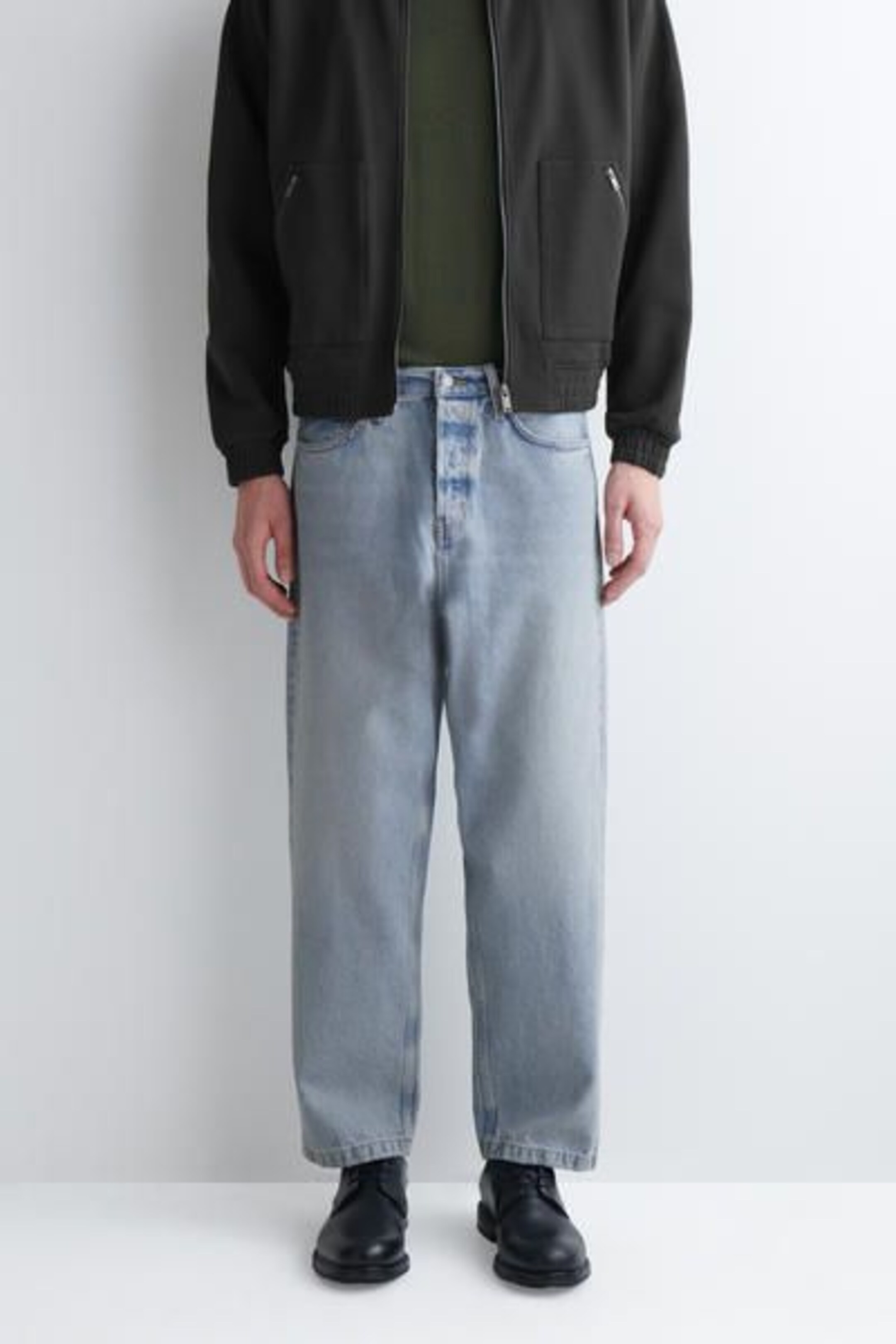Person wearing light blue jeans, a dark jacket, and black shoes, standing against a white background.