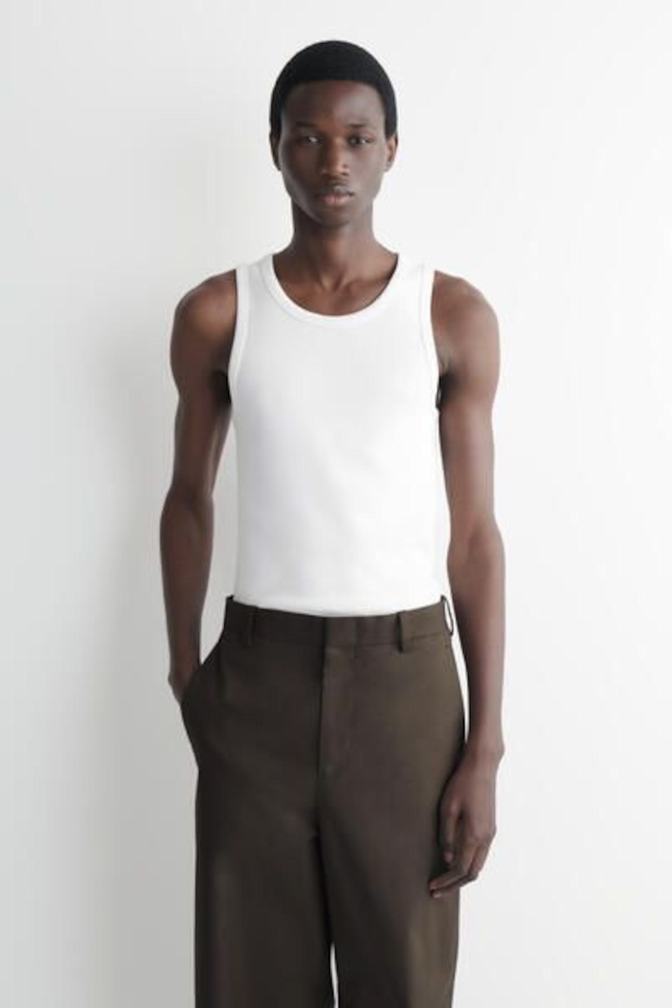 A person wearing a white sleeveless top and dark brown trousers, standing against a plain white background.