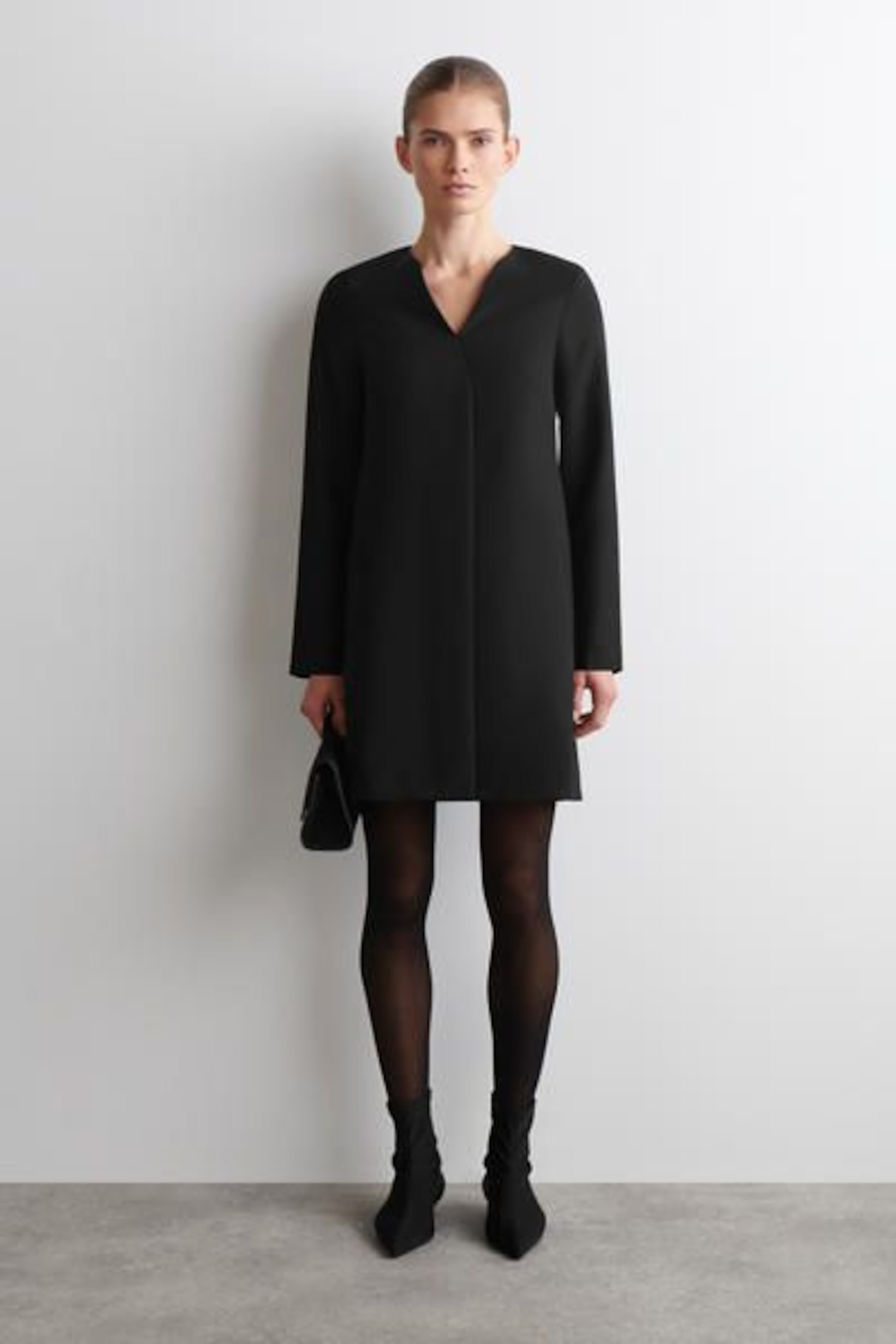 A person stands in a minimalist setting wearing a black dress, black tights, and holding a small black handbag, with hair tied back.