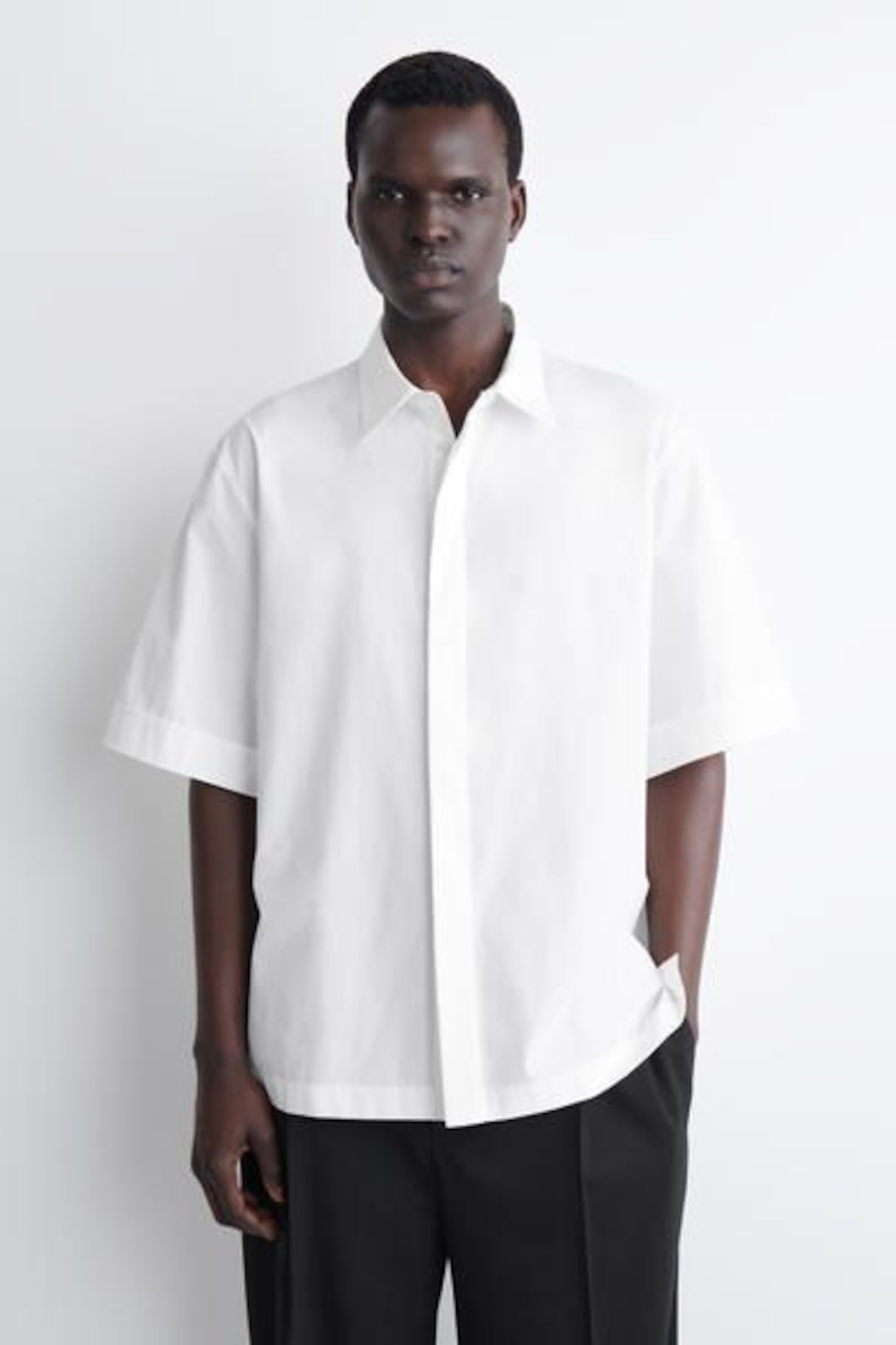 A person wearing a short-sleeved white button-up shirt and black pants, standing against a plain white background.
