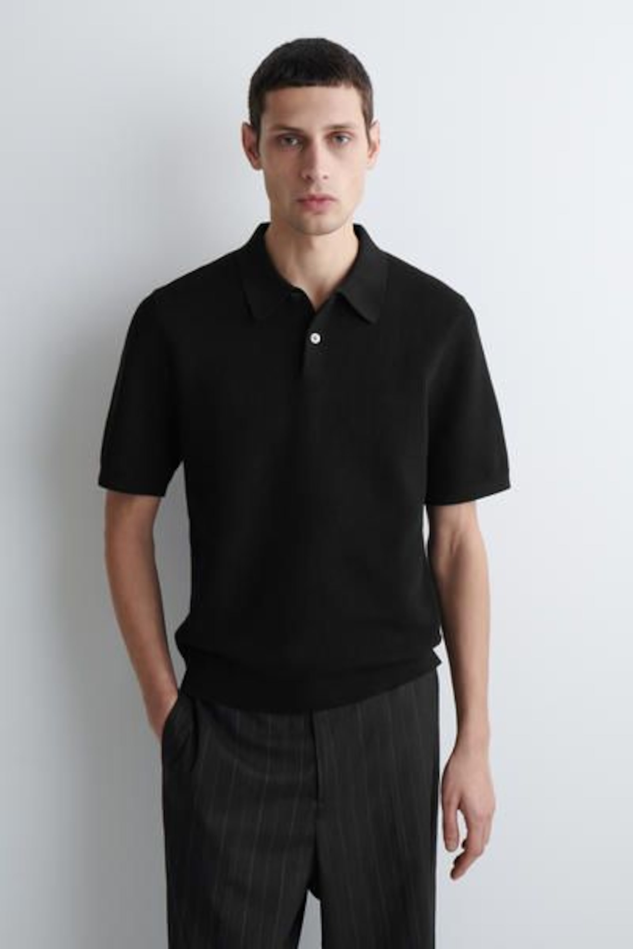 A person wearing a black short-sleeve polo shirt and dark pinstriped trousers stands against a light background with hands in pockets.
