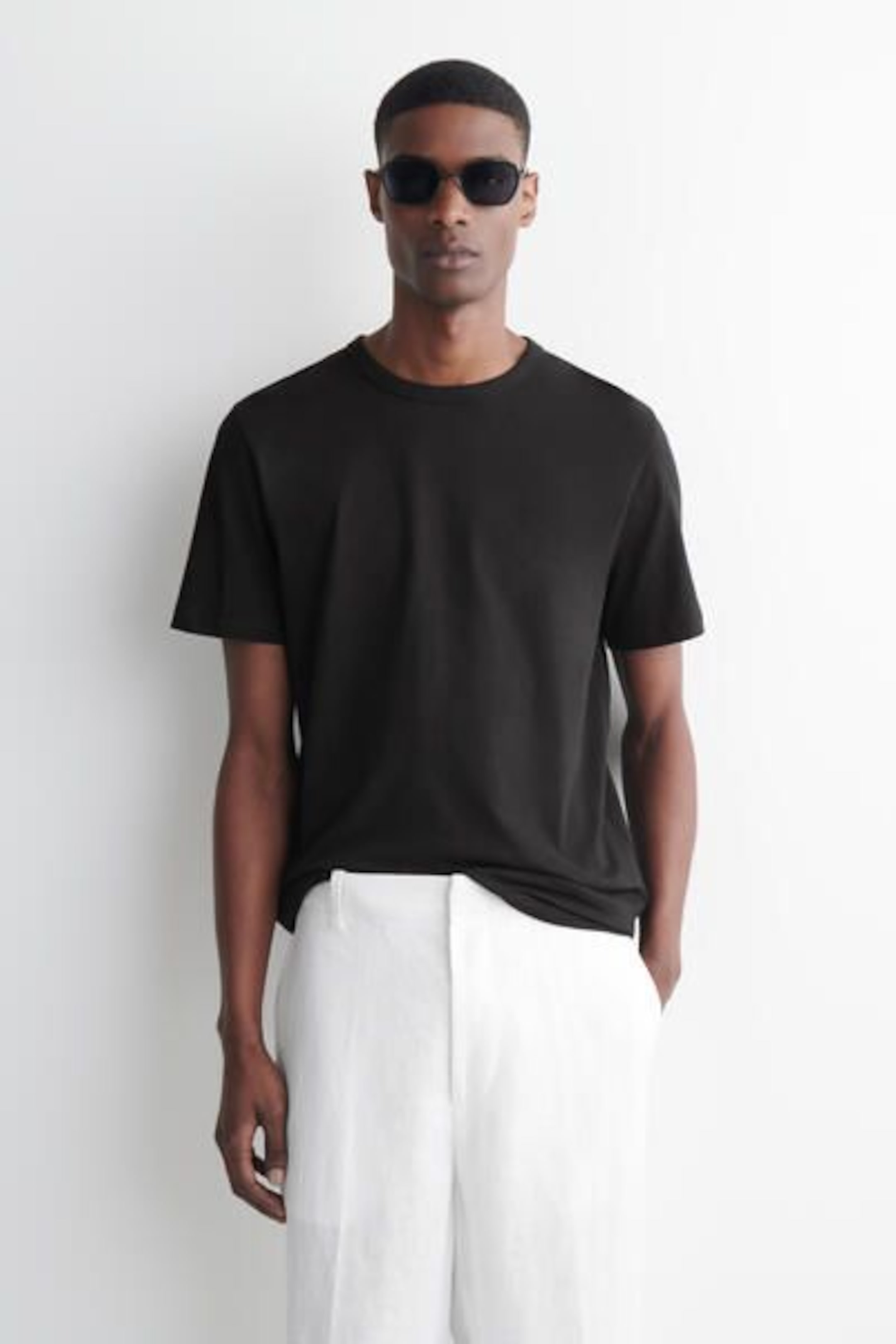 Man wearing sunglasses, a black t-shirt, and white pants stands against a plain white background.