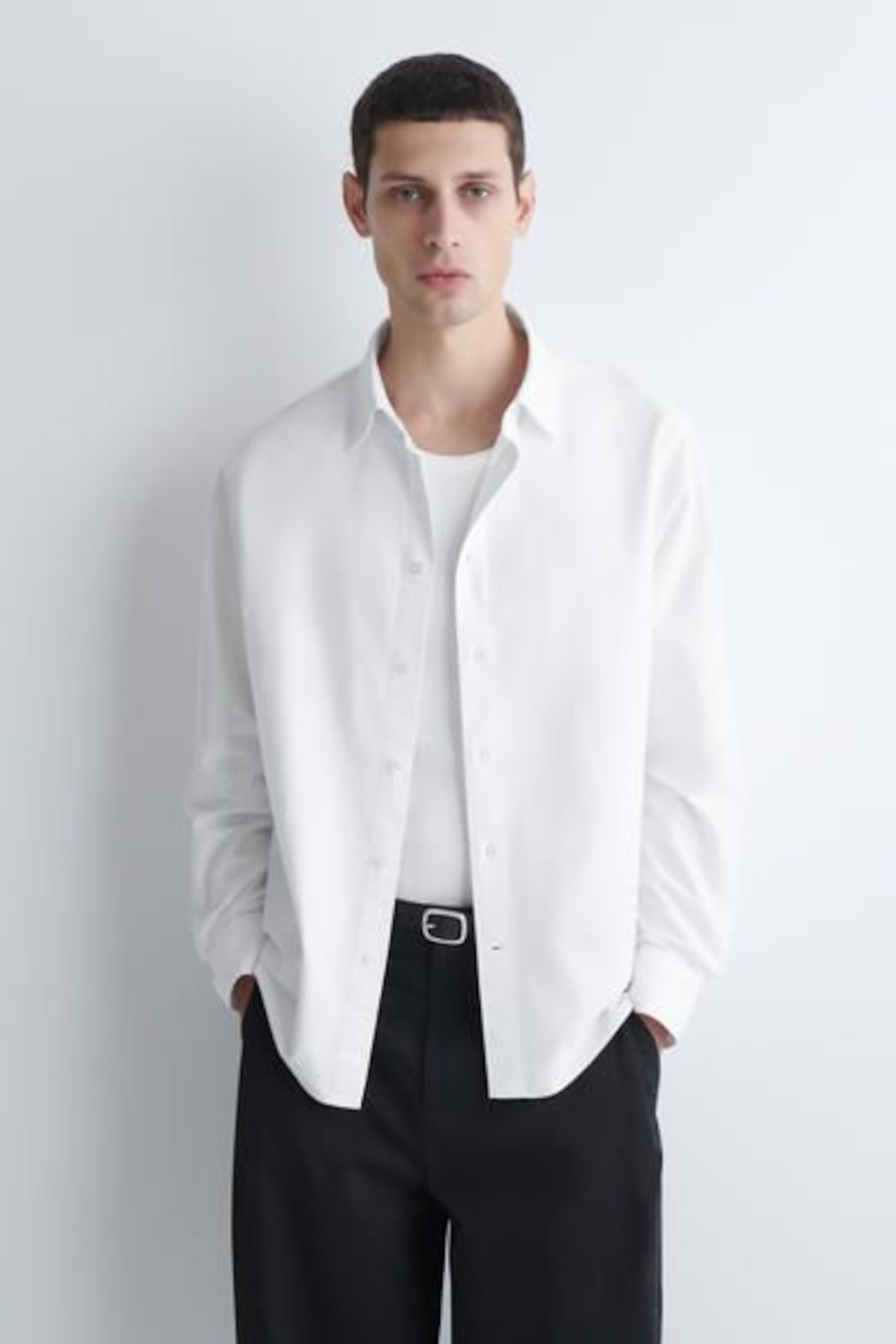 A man wearing a white button-up shirt over a white t-shirt and black pants stands against a white background.