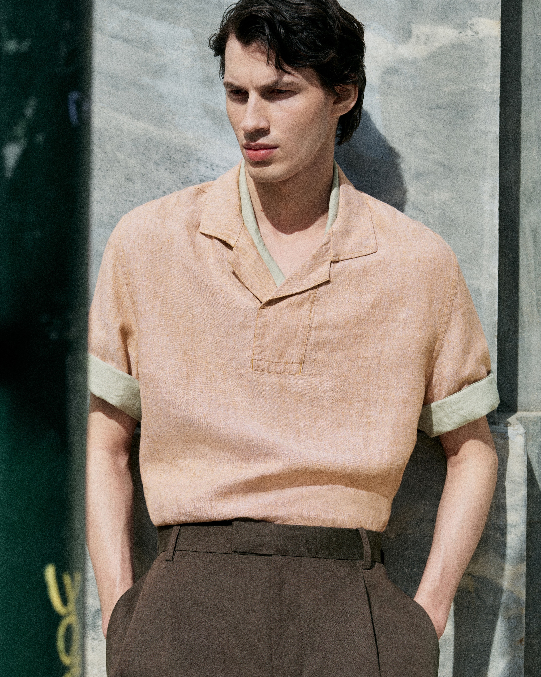 Person in a peach shirt and brown pants leaning against a stone wall, gazing sideways in soft sunlight.