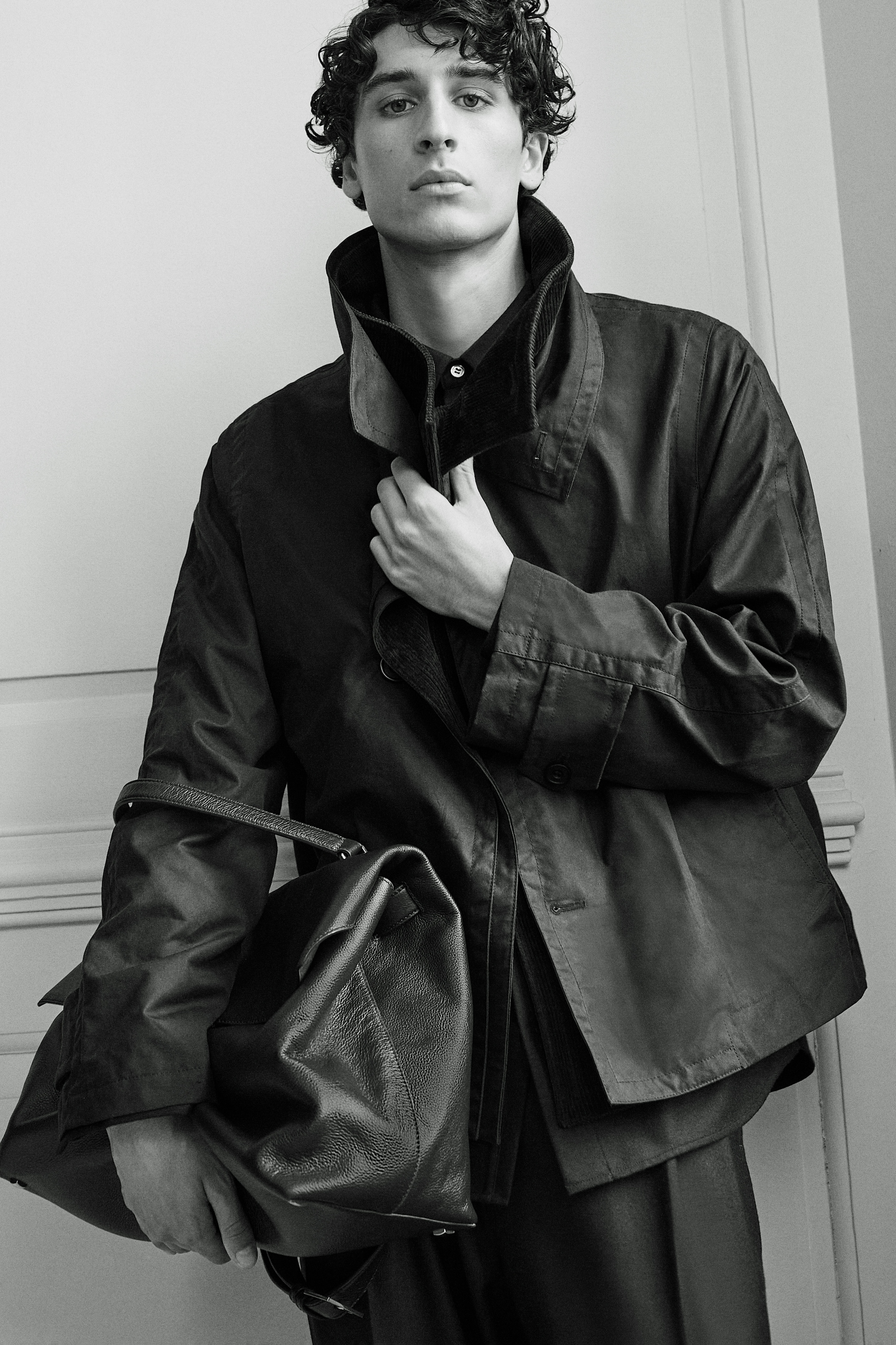 A person with curly hair wearing a high-collared jacket and holding a large leather bag, posed confidently against a plain background.