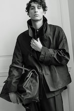 Placeholder for: A person with curly hair wearing a high-collared jacket and holding a large leather bag, posed confidently against a plain background.