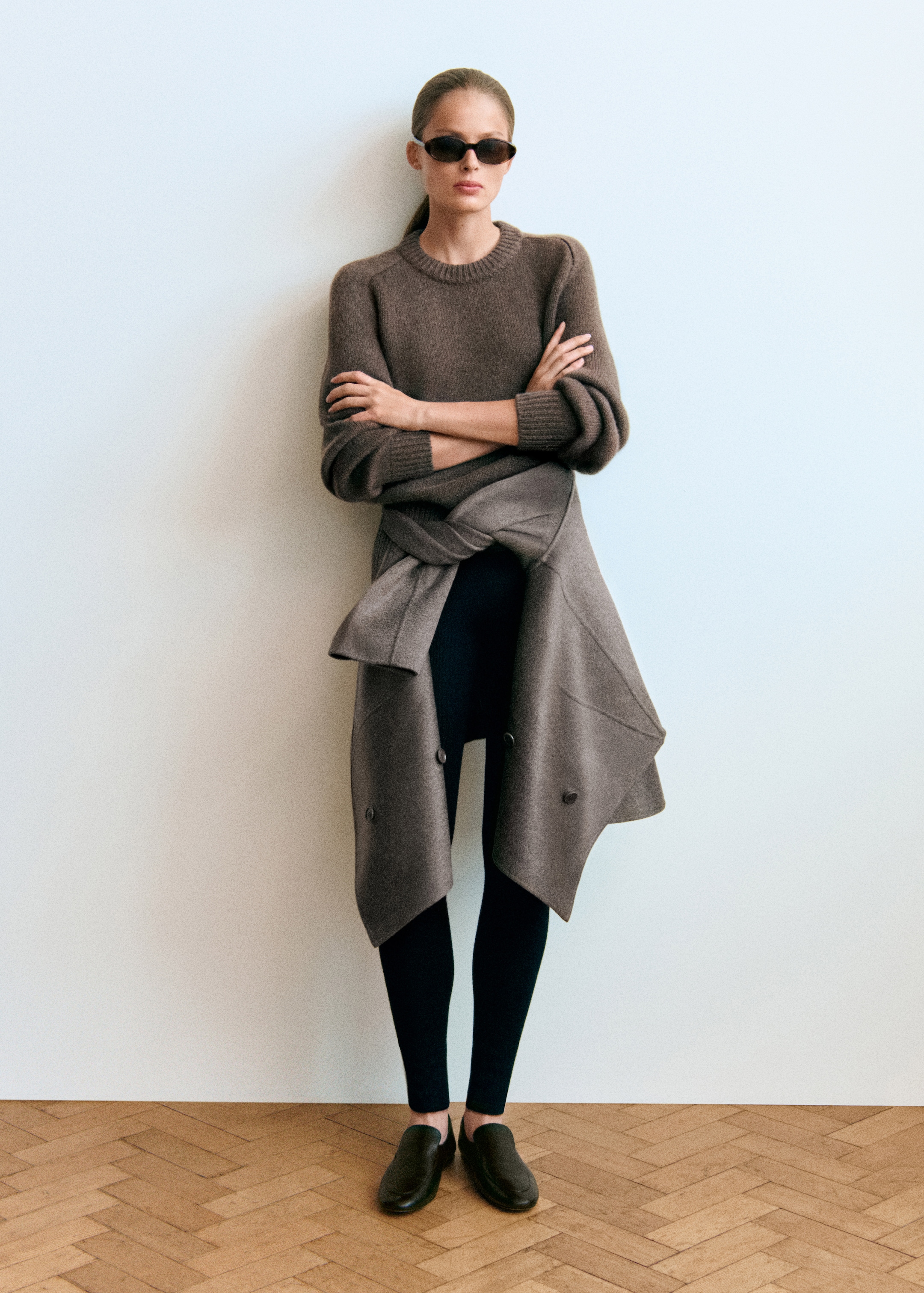 Person wearing sunglasses, a brown sweater, dark leggings, brown loafers, and a gray coat tied around the waist, standing against a white wall.