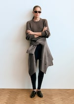 Placeholder for: Person wearing sunglasses, a brown sweater, dark leggings, brown loafers, and a gray coat tied around the waist, standing against a white wall.