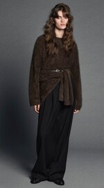 Placeholder for: A person with long hair wearing a brown textured sweater with a tie belt and black wide-leg pants, standing against a gray background.
