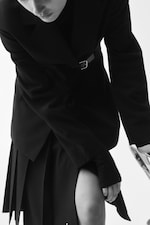 Placeholder for: Black and white image of a person in a stylish black coat and skirt, leaning forward, holding a transparent umbrella.