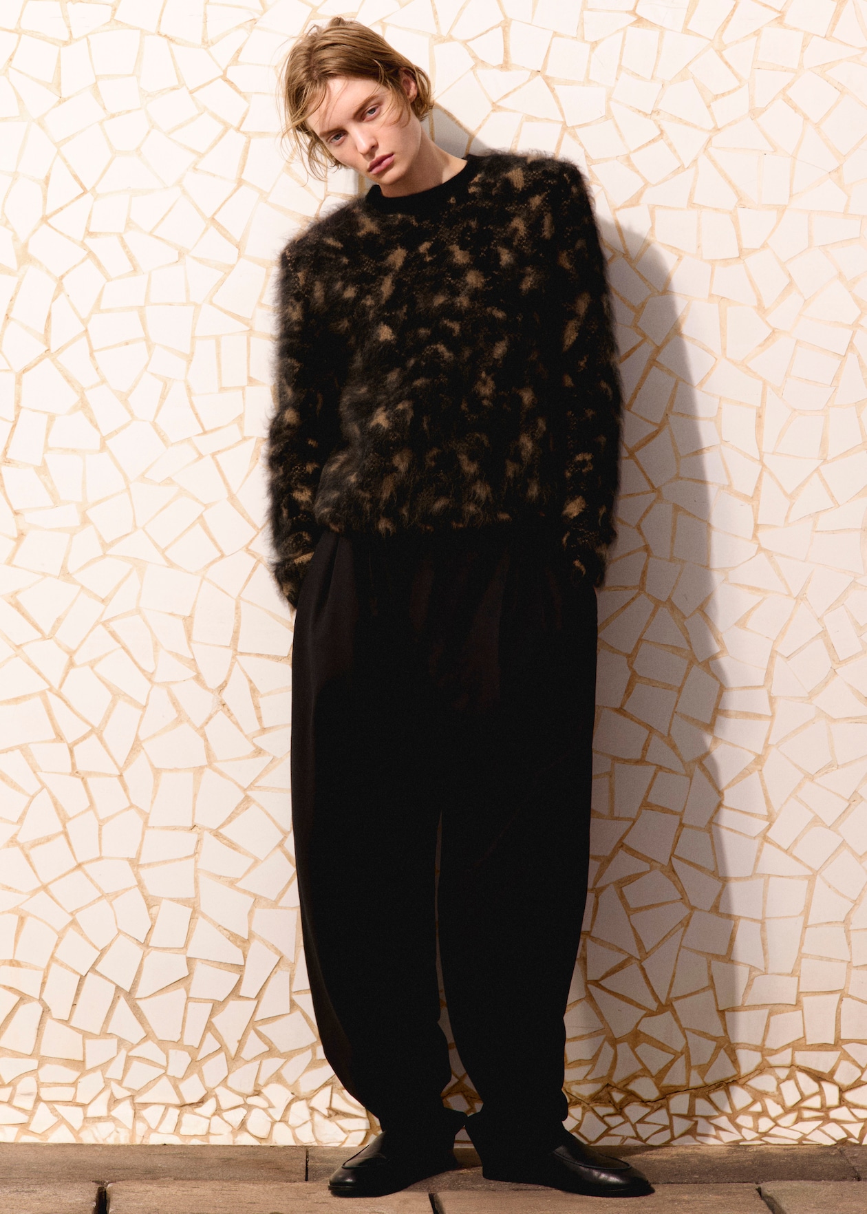 Person wearing a fuzzy, dark-patterned sweater and loose black pants stands against a white mosaic wall, looking relaxed.