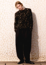 Placeholder for: Person with tousled hair in a fuzzy sweater and loose pants leans against a mosaic wall, casting a shadow.