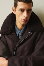 Placeholder for: A person wearing a dark brown coat with a fleece collar and a gray shirt underneath, looking slightly to the side.