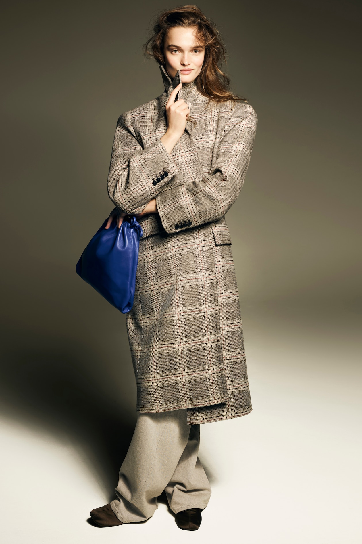 A person in a plaid coat poses confidently, holding a blue bag, with one hand on their chin, against a neutral background.