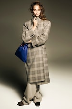 Placeholder for: A person in a plaid coat poses confidently, holding a blue bag, with one hand on their chin, against a neutral background.