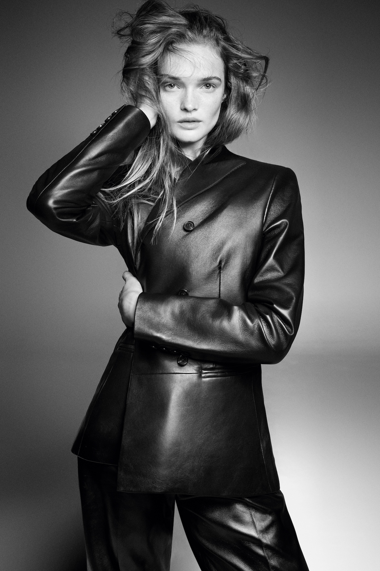 A person in a black leather jacket and pants poses confidently, their hair tousled, with a hand touching their head against a gray background.