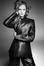 Placeholder for: A person in a black leather jacket and pants poses confidently, their hair tousled, with a hand touching their head against a gray background.