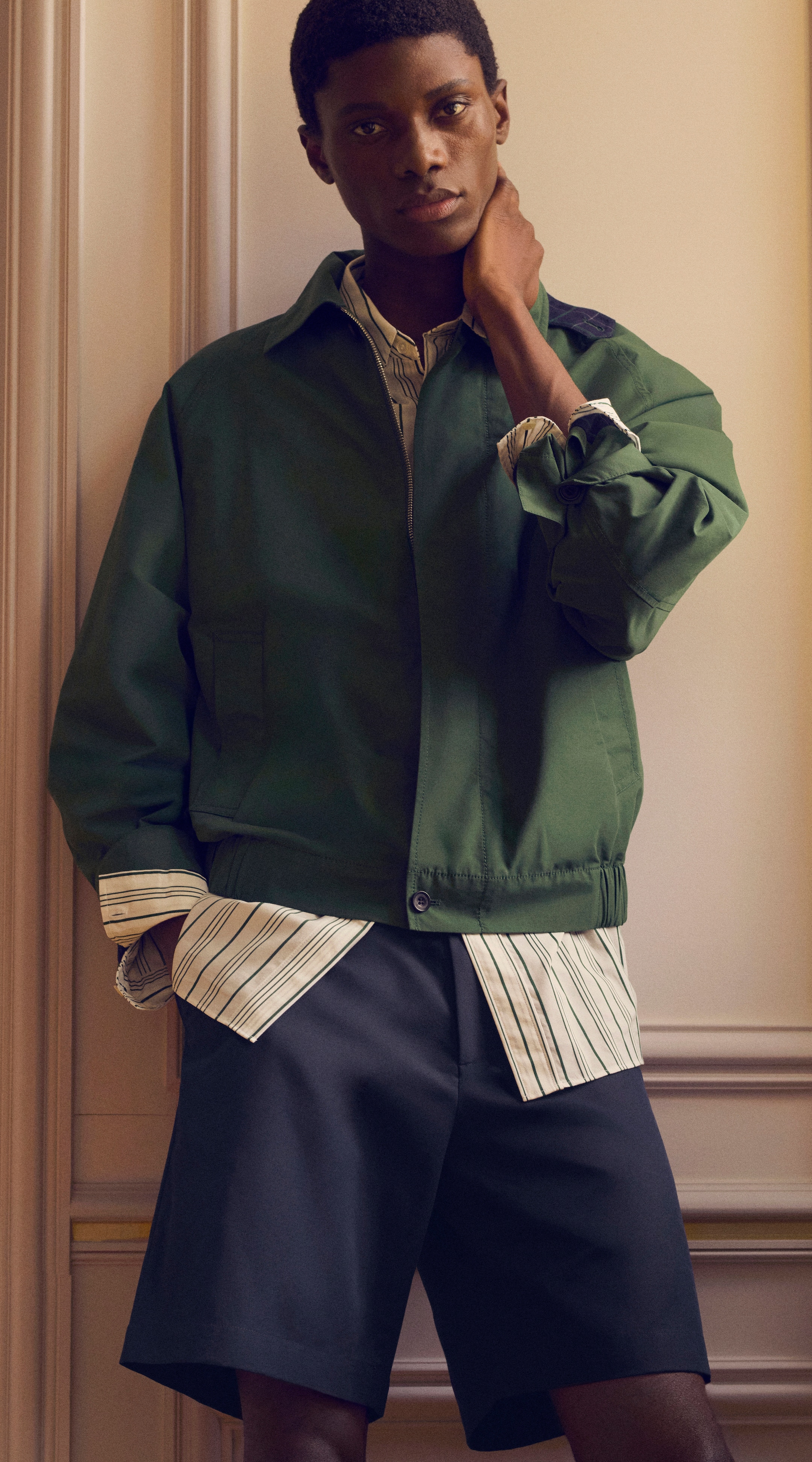 Person wearing a green jacket over a striped shirt and dark shorts, posing with one hand on their neck, standing in front of a paneled wall.
