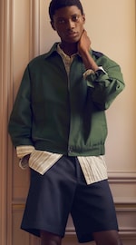 Placeholder for: Person wearing a green jacket over a striped shirt and dark shorts, posing with one hand on their neck, standing in front of a paneled wall.