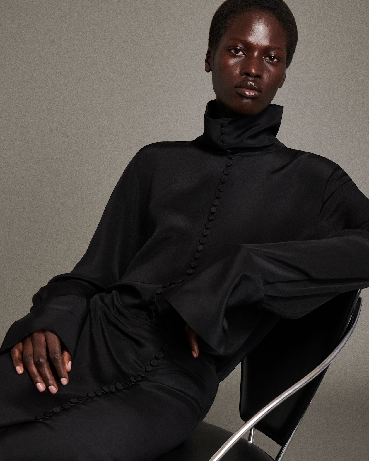 Person sitting in a chair wearing a high-collared black dress with decorative buttons, with a neutral background.