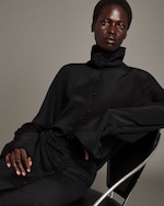 Placeholder for: Person sitting in a chair wearing a high-collared black dress with decorative buttons, with a neutral background.