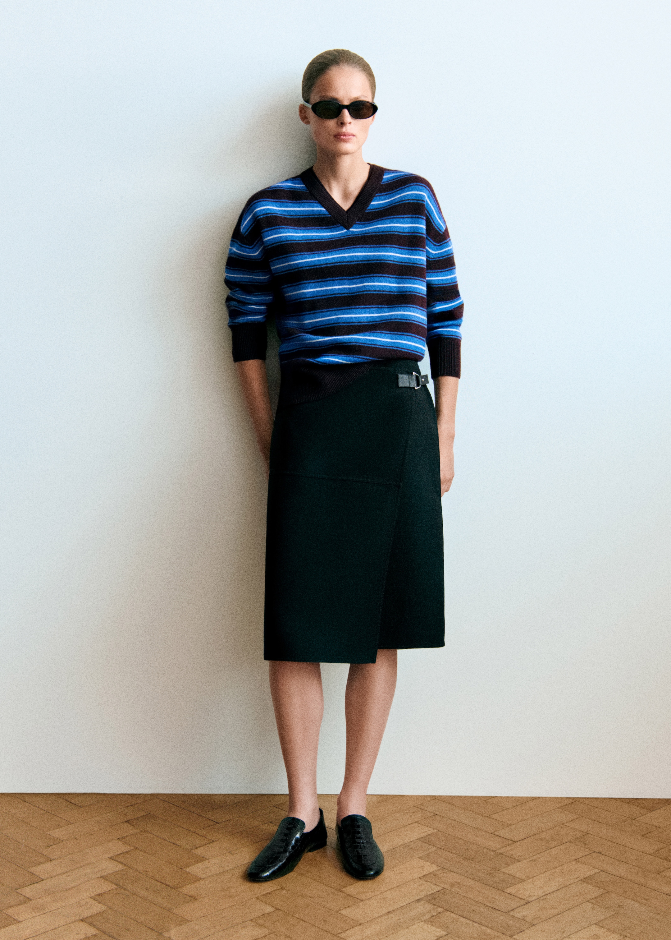 A person wearing sunglasses, a striped sweater, and a black skirt stands against a white wall on a wooden floor.