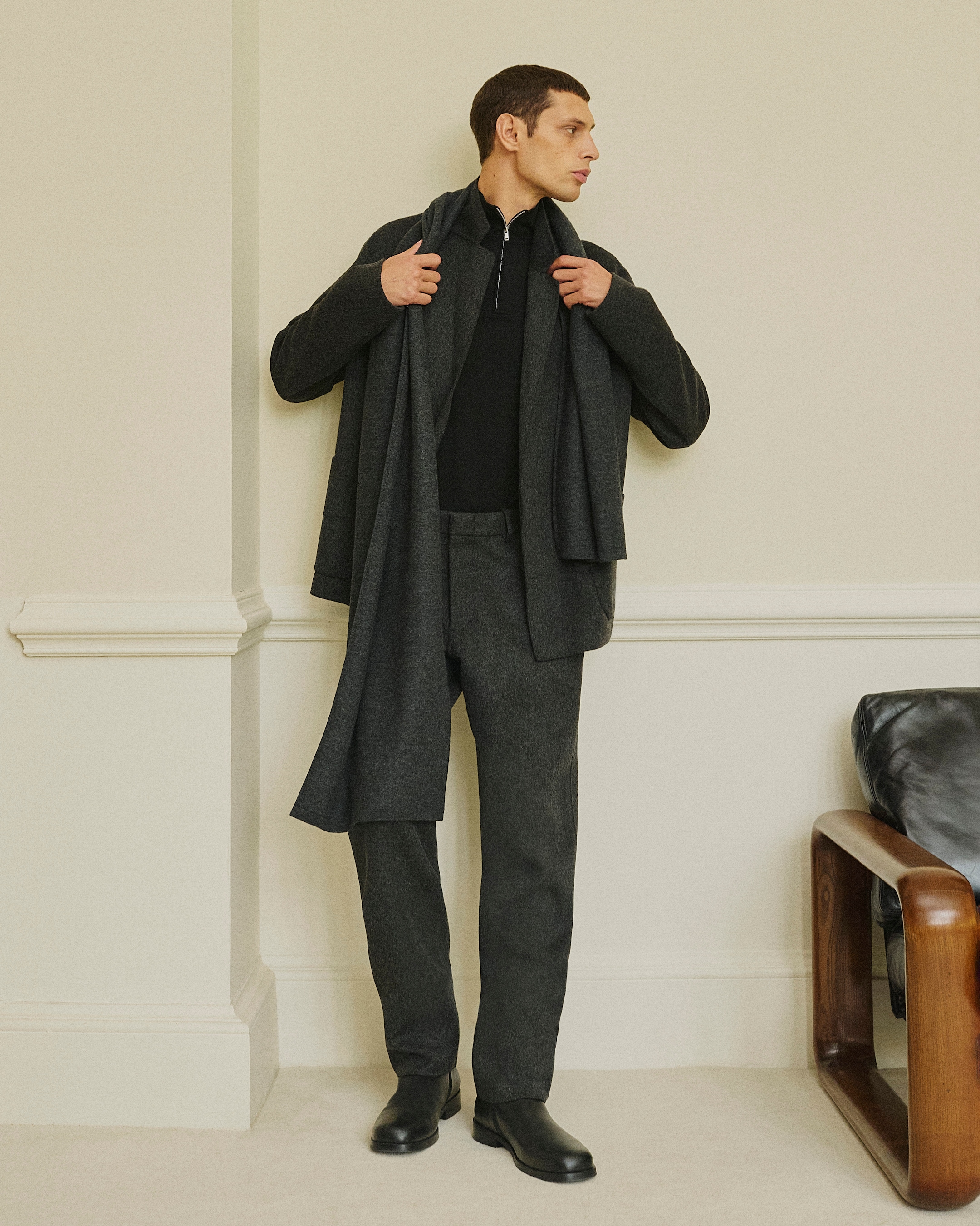 A man wearing a black zip-up shirt, gray jacket, and matching pants with a scarf, stands indoors next to a cream wall, looking to his left.