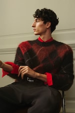 Placeholder for: A person with curly hair wearing a red and brown plaid sweater over a red shirt sits in a chair, looking to the side.