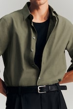 Placeholder for: Man wearing a green button-up shirt over a black t-shirt, with black trousers and a belt.