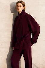 Placeholder for: Person in a burgundy coat and pants stands against a light-colored, textured wall, looking to the side.