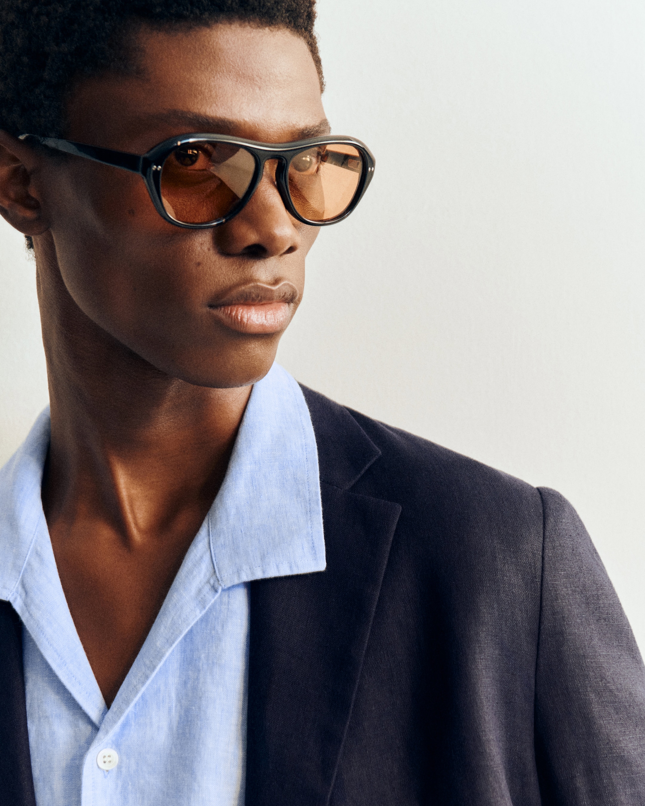 Person wearing brown-tinted glasses, a light blue shirt, and a dark blazer, gazing to the side against a plain background.
