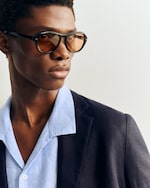 Placeholder for: Person wearing brown-tinted glasses, a light blue shirt, and a dark blazer, gazing to the side against a plain background.