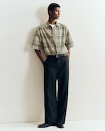 Placeholder for: Person in a plaid short-sleeve shirt, white tee, and black wide-leg pants stands against a white wall with hands in pockets.