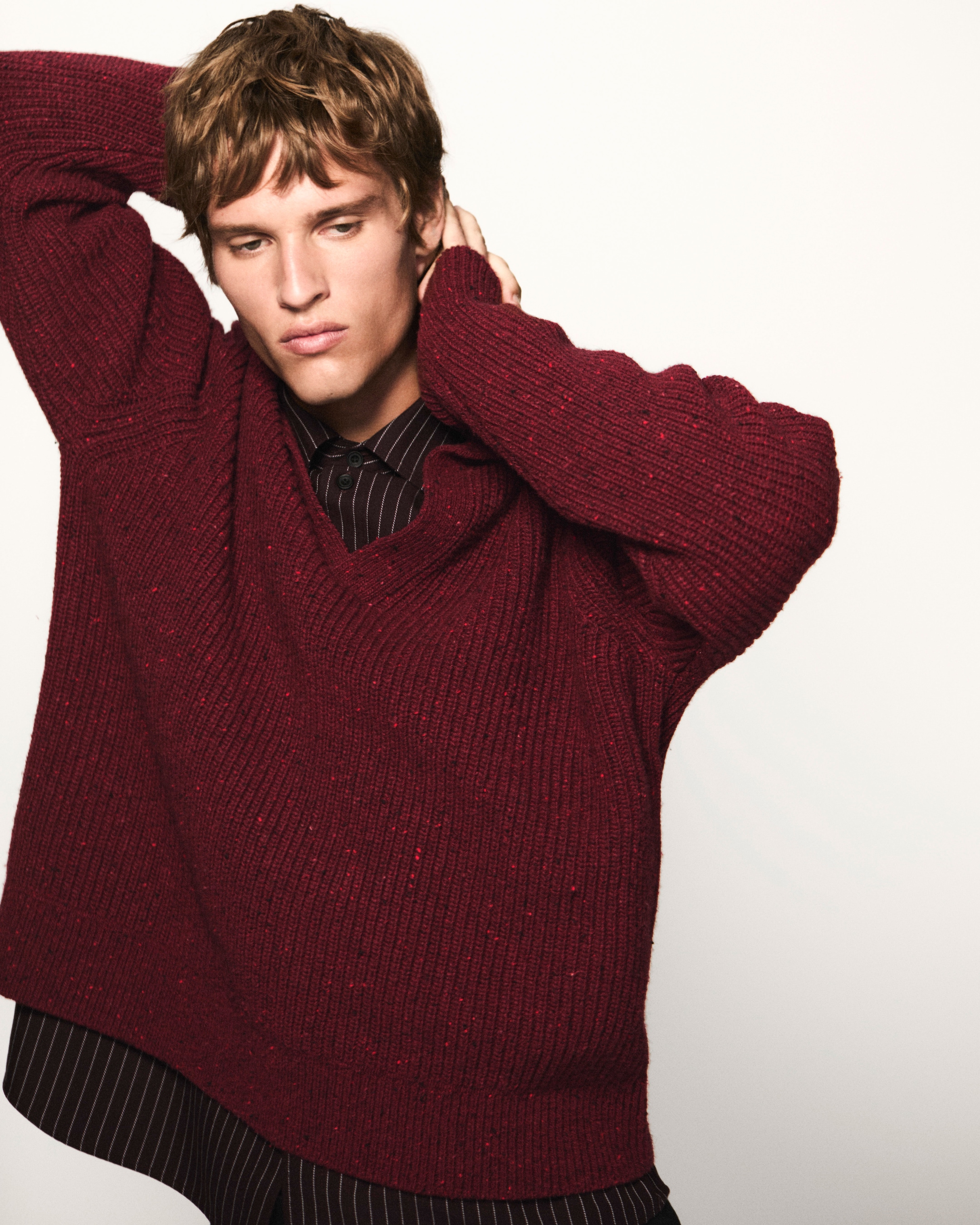 Person in a red sweater over a striped shirt poses with arms raised against a plain background.