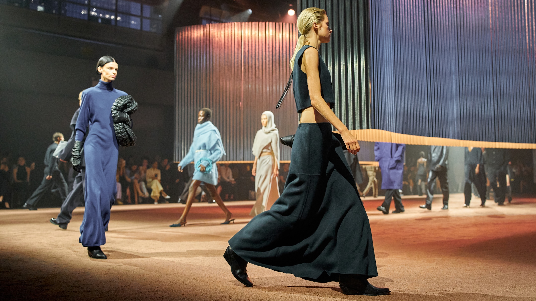 Models walk a runway in elegant outfits, featuring long dresses and layered garments, under soft lighting in a stylish, industrial setting.