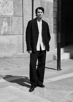 Placeholder for: Man in black suit and white shirt stands on a city street, leaning on a wall, in a black and white photograph.